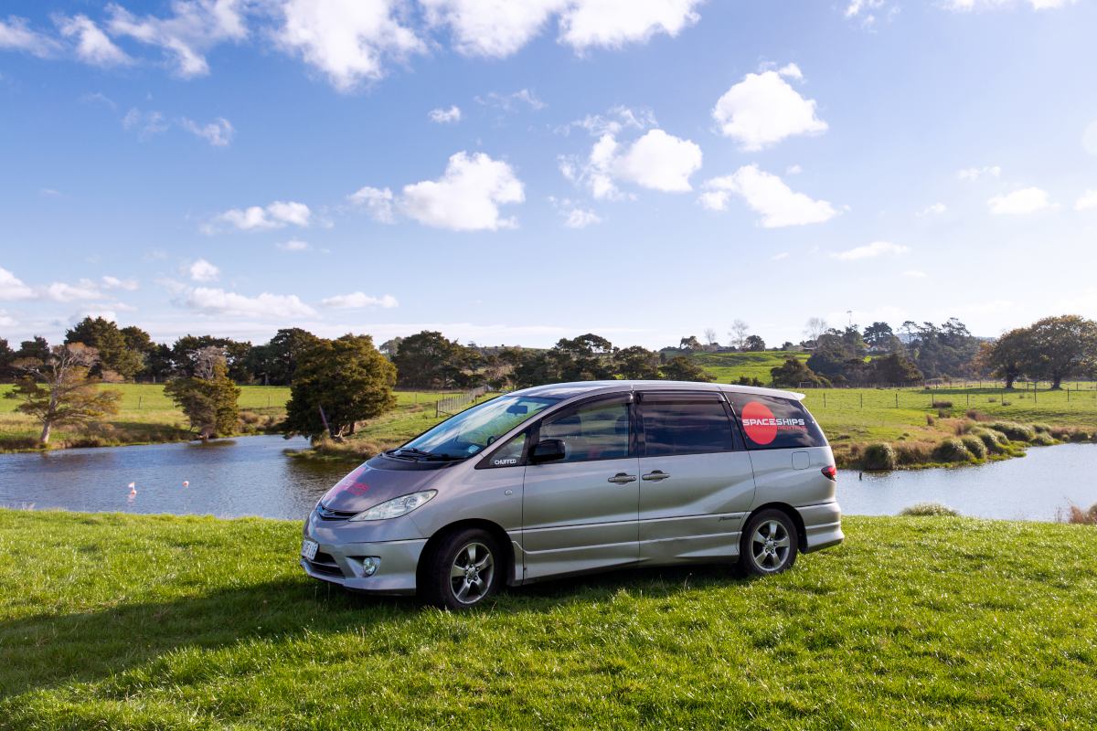 Silver campervan parked parked on the grass in front of a small pond with rolling rural hills in the background