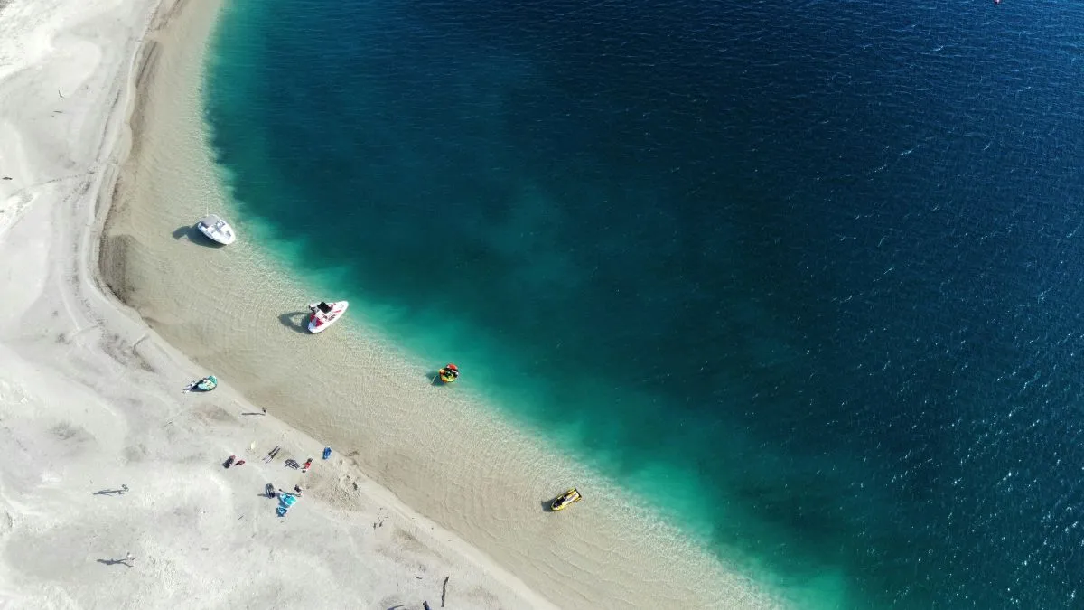 boats and people at the side of a clear blue lake with white sand
