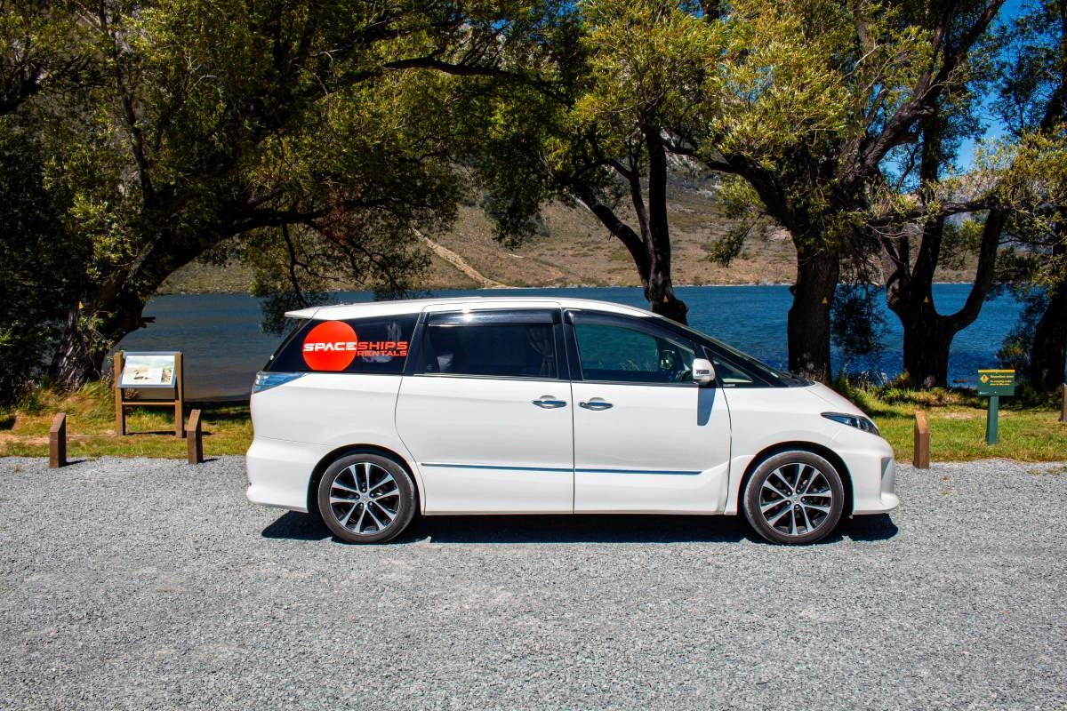 Side view of a white campervan parked in front of a lake with large trees in the background