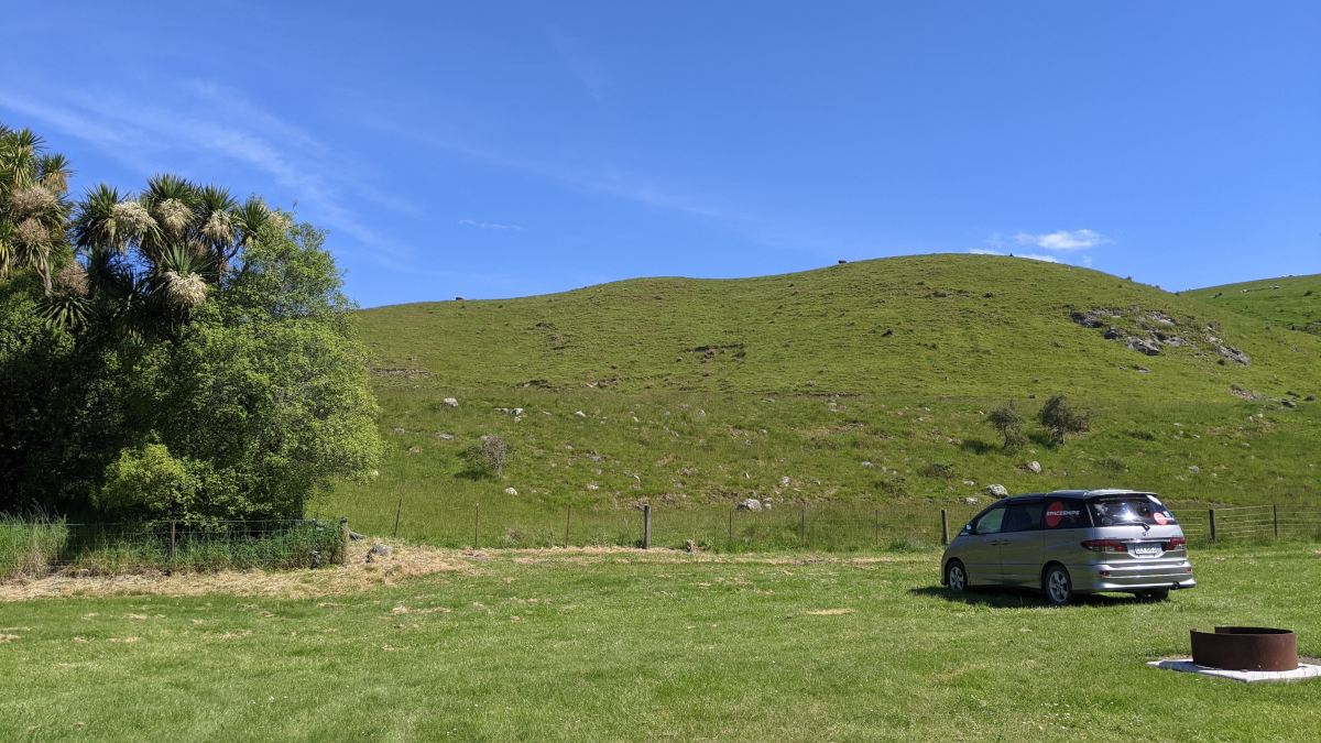 Green grass and green hills with a Spaceships campervan parked in front of it. Sunny day at Waihi Gorge campsite